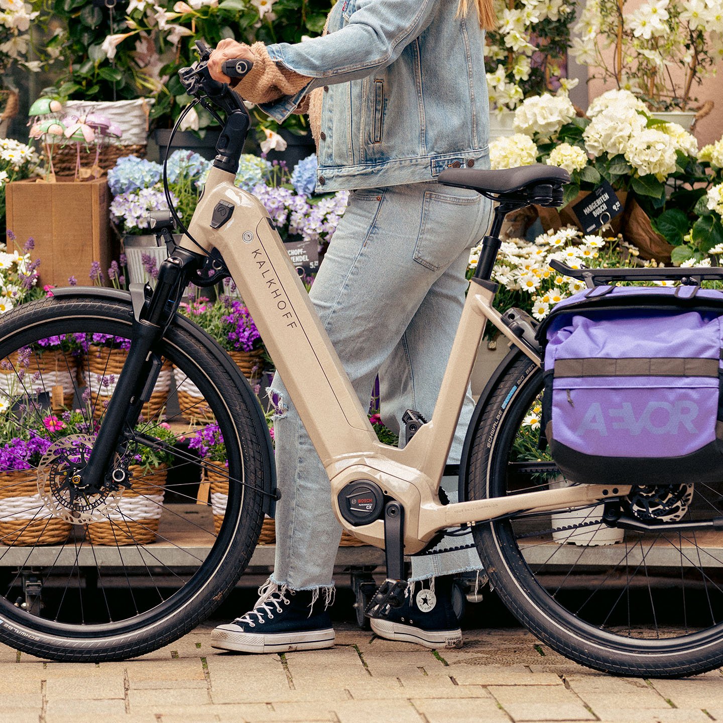 Eine Person steht neben einem beigen Fahrrad mit einer violetten Tasche vor einem Blumenladen mit verschiedenen Pflanzen.
