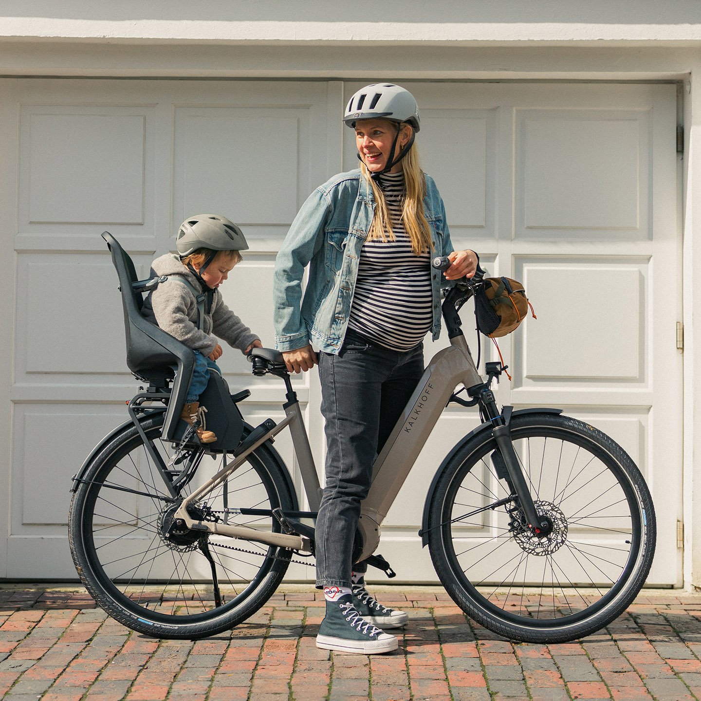 Frau mit Helm und Kind auf dem Fahrradsitz, vor einem Garagentor stehend. die Frau lächelt und hält das Fahrrad fest.