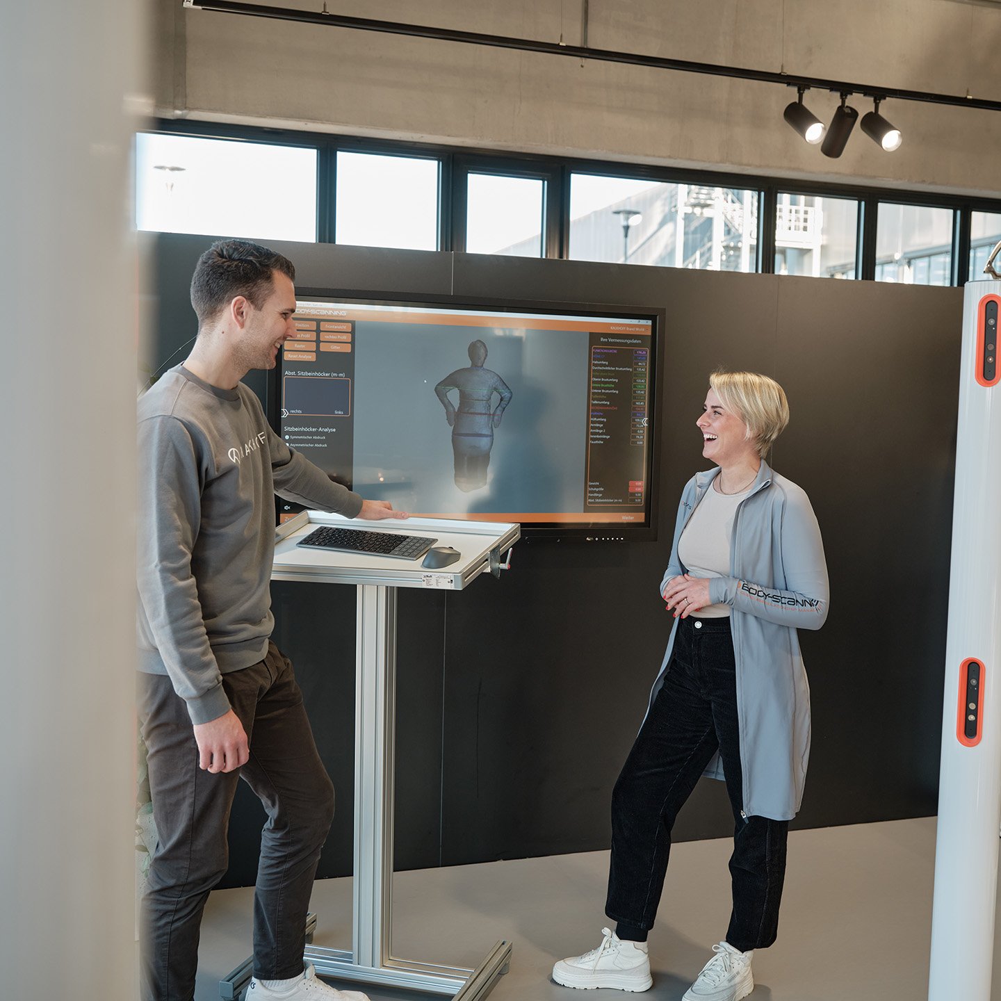 Zwei Personen stehen vor einem Bildschirm, auf dem ein 3D-Körperscan angezeigt wird.