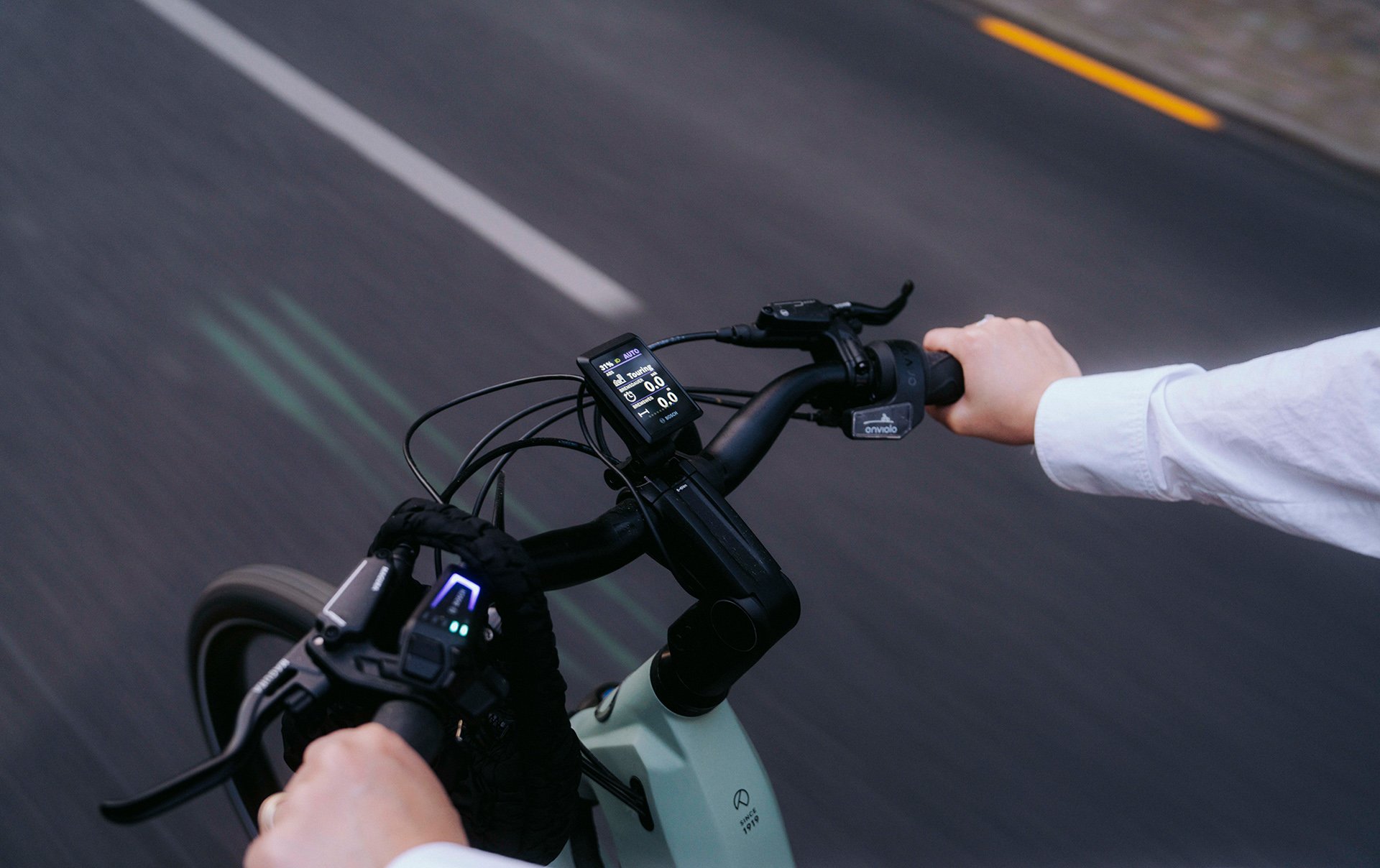 Person riding an e-bike on a road, hands on handlebars with a digital display showing speed and battery level, motion blur in the background.