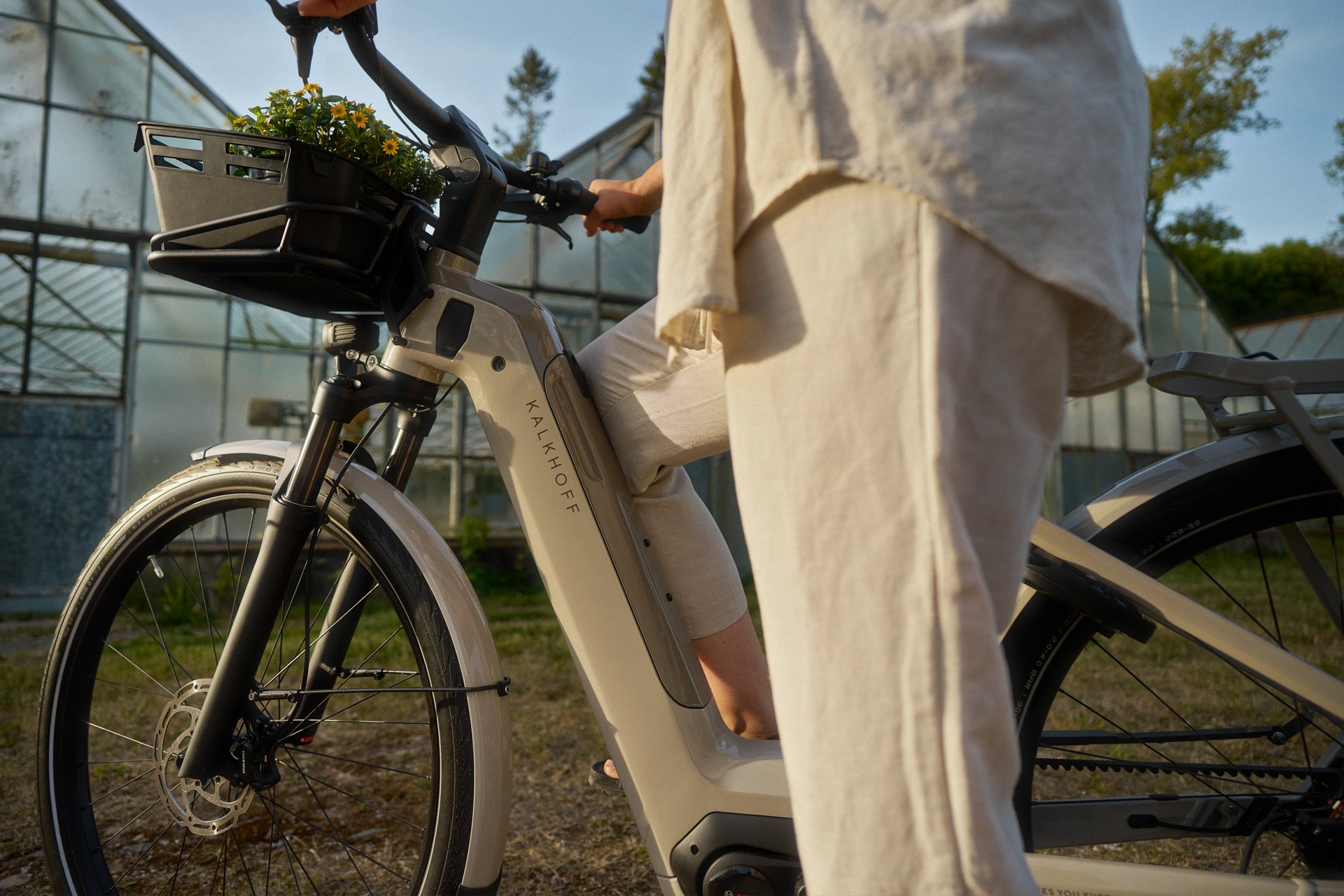 Person in light clothing stands next to an electric bike with a basket of flowers, near greenhouses in a sunny outdoor setting.