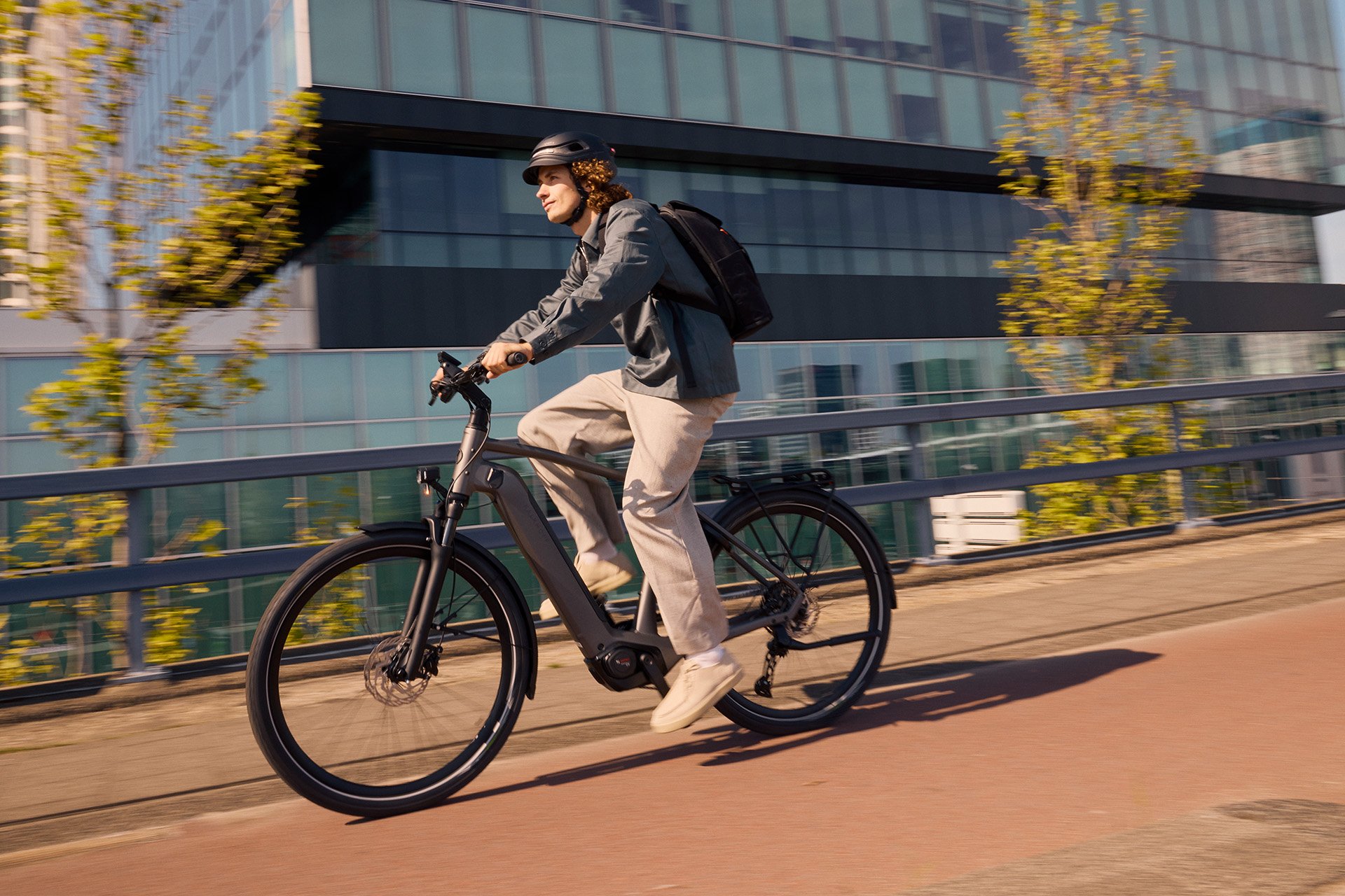 Zusätzlich zur mechanischen Entkopplung sorgt die Technik im Motor dafür, dass die Tretunterstützung sofort beendet wird, sobald du die Pedalbewegung stoppst. Person riding an electric bike on a city path, wearing a helmet and backpack, with modern glass buildings in the background.