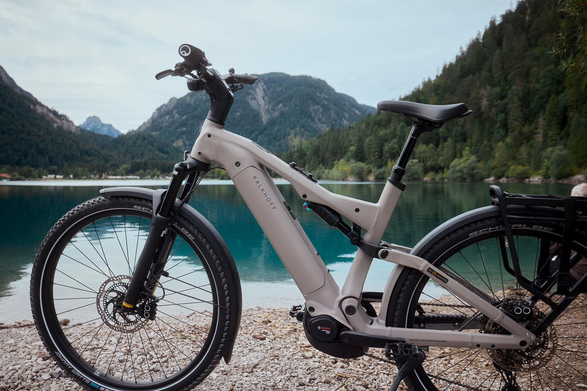 A white electric bike is parked on a pebbled shore, overlooking a calm blue lake surrounded by lush green mountains under a cloudy sky.