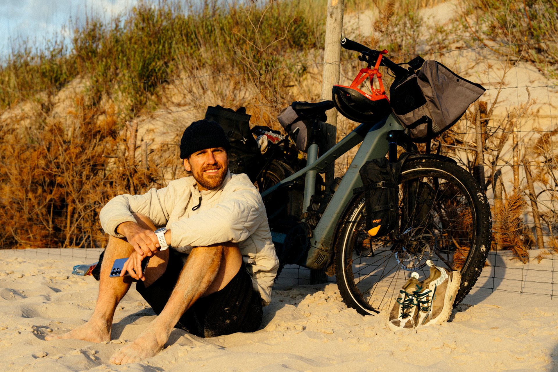 Man sitting on sandy beach next to a loaded bicycle, wearing a beanie and light jacket, with a thoughtful expression.