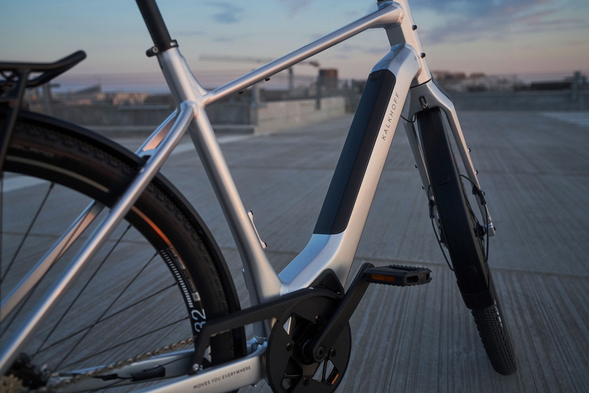 Close-up of a sleek silver e-bike frame on a rooftop, showcasing its integrated battery and modern design against a twilight sky.