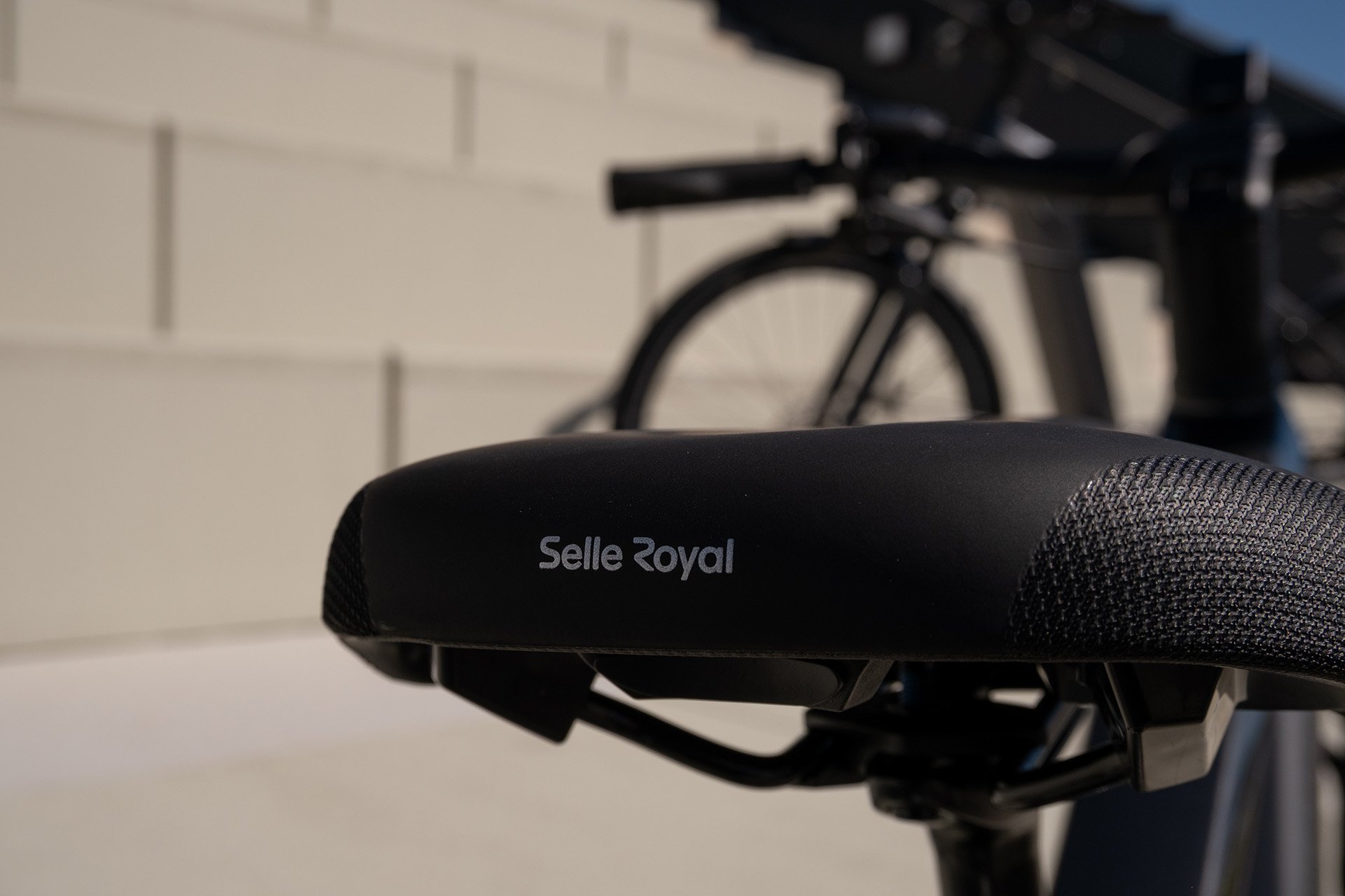 Der Sattel ist für den Fahrkomfort auf dem E-Bike essenziell. Close-up of a Selle Royal bike saddle with a blurred black bike in the background, set against a light-tiled wall.
