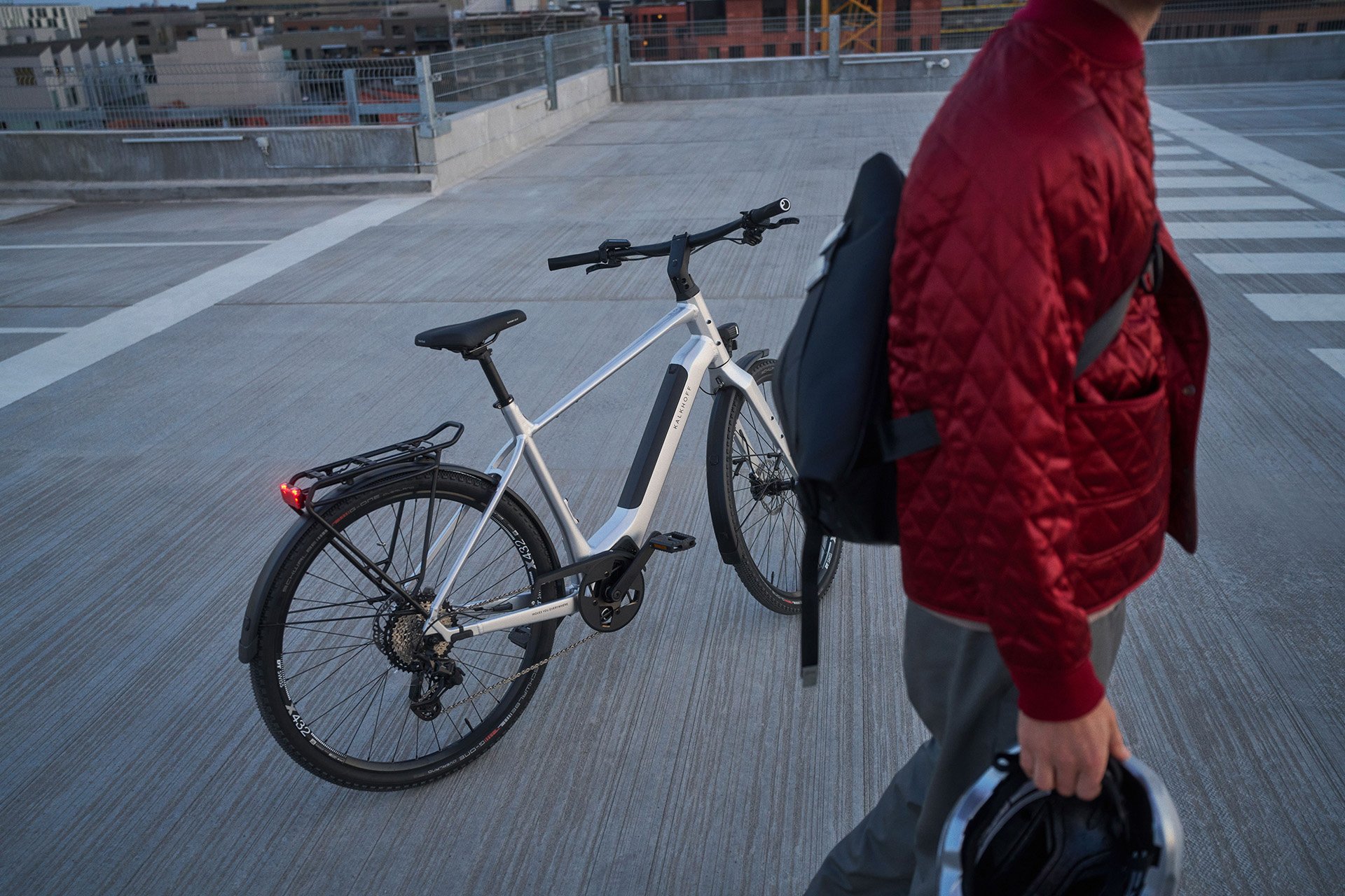 Person in red jacket holding a helmet, walks on rooftop next to a white electric bicycle with a rear rack.