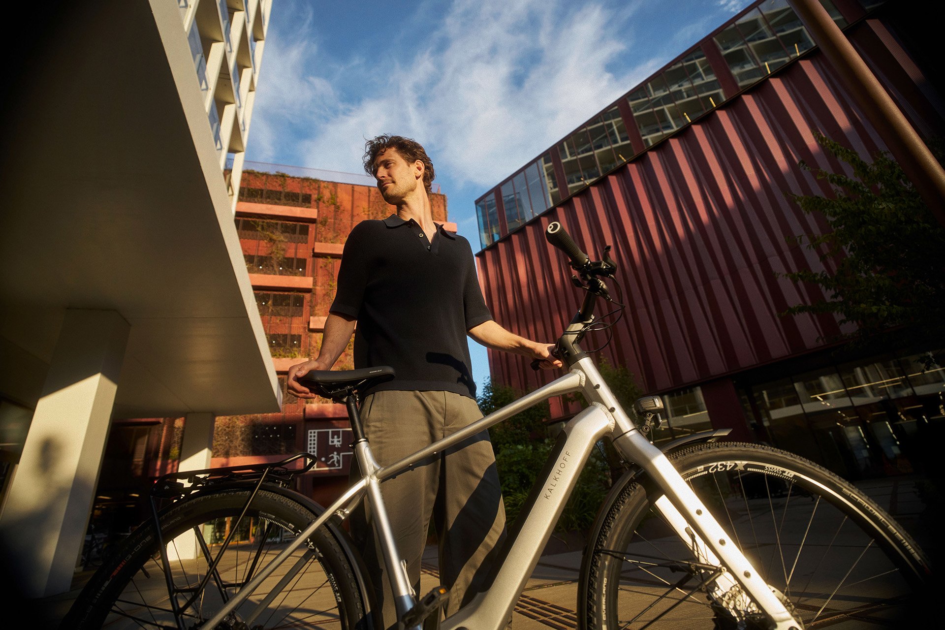 Man stands with a bicycle in an urban setting, surrounded by modern buildings under a clear blue sky.