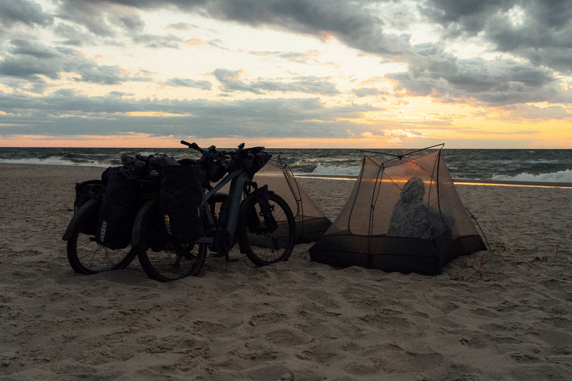 Two bicycles and small tents on a sandy beach at sunset, with a cloudy sky and ocean in the background.