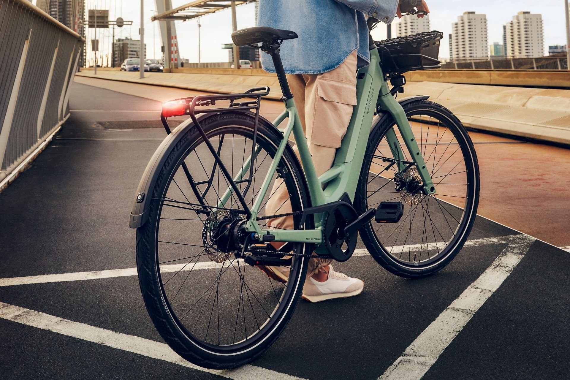 Kalkhoff E-Bike Eine Frau hält ein Kalkhoff Light Modell auf einer Straßenbrücke, es ist nur der Unterkörper der Frau zu sehen. Das E-Bike wurde seitlich von hinten fotografiert.