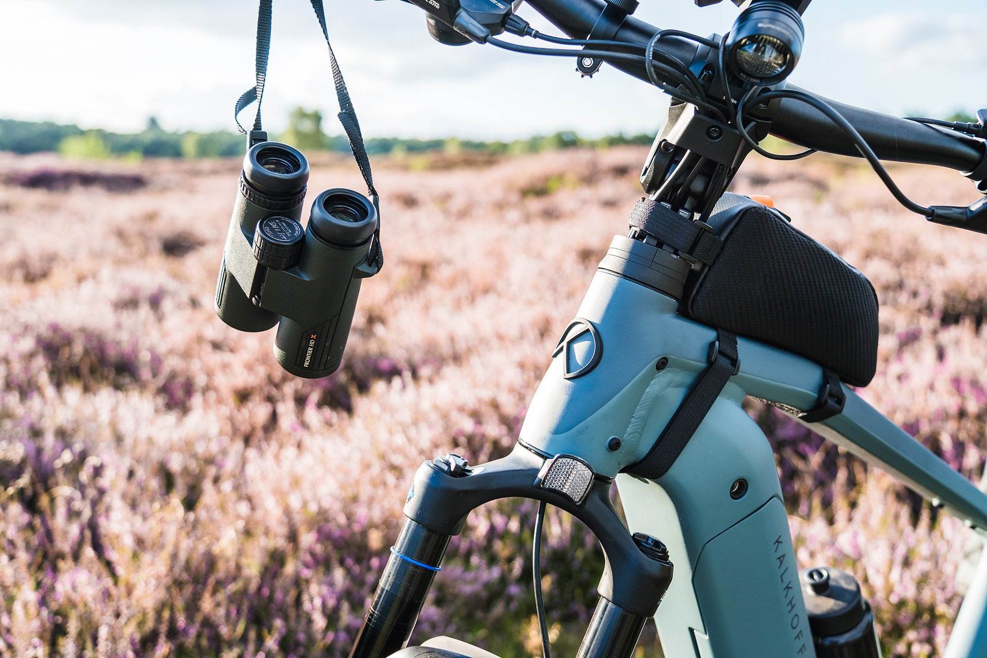 Binoculars hang from a mountain bike's handlebars, with a field of purple flowers and green trees in the background under a cloudy sky.