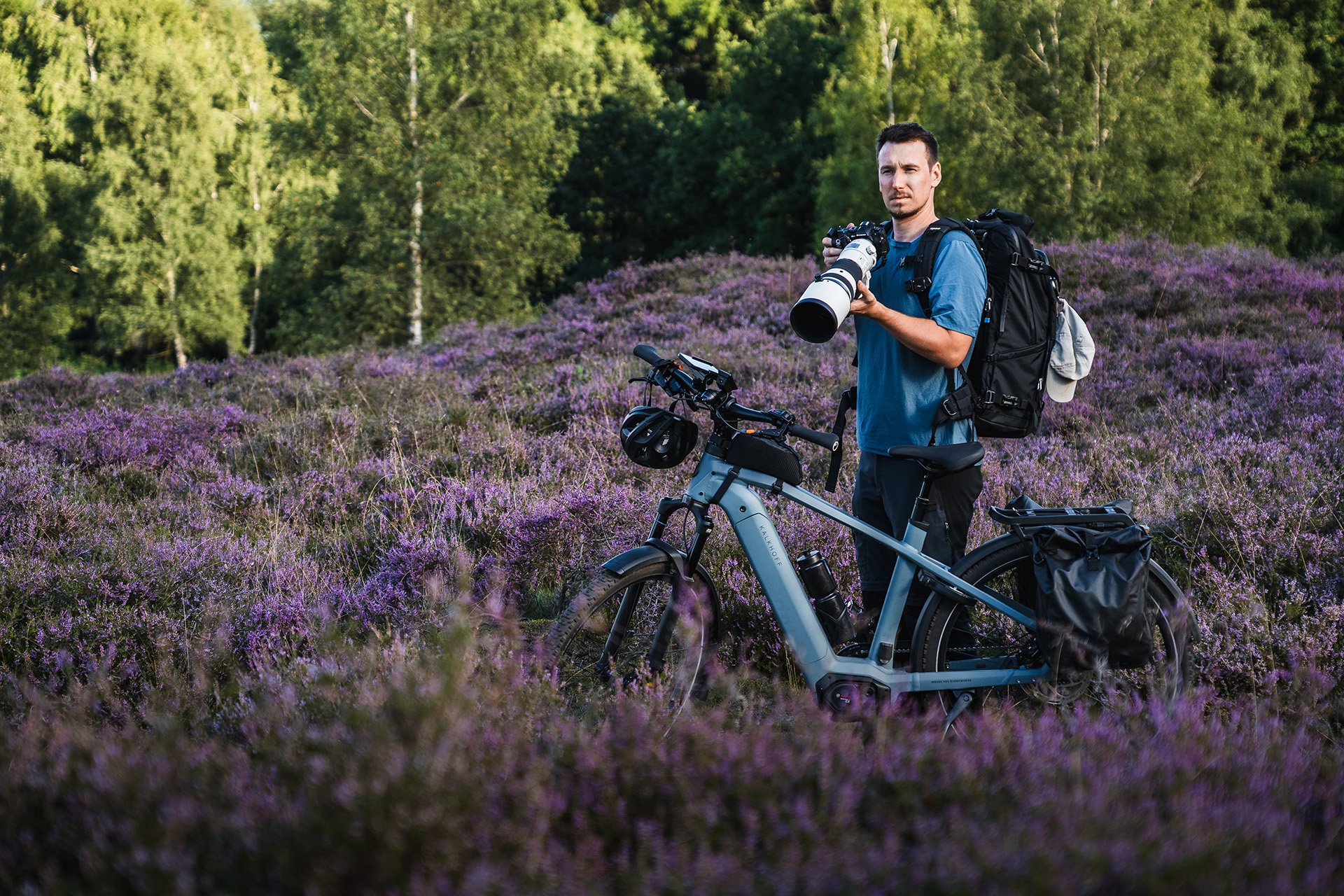 A man stands beside his bicycle in a field of purple flowers, holding a camera with a large lens, surrounded by green trees.