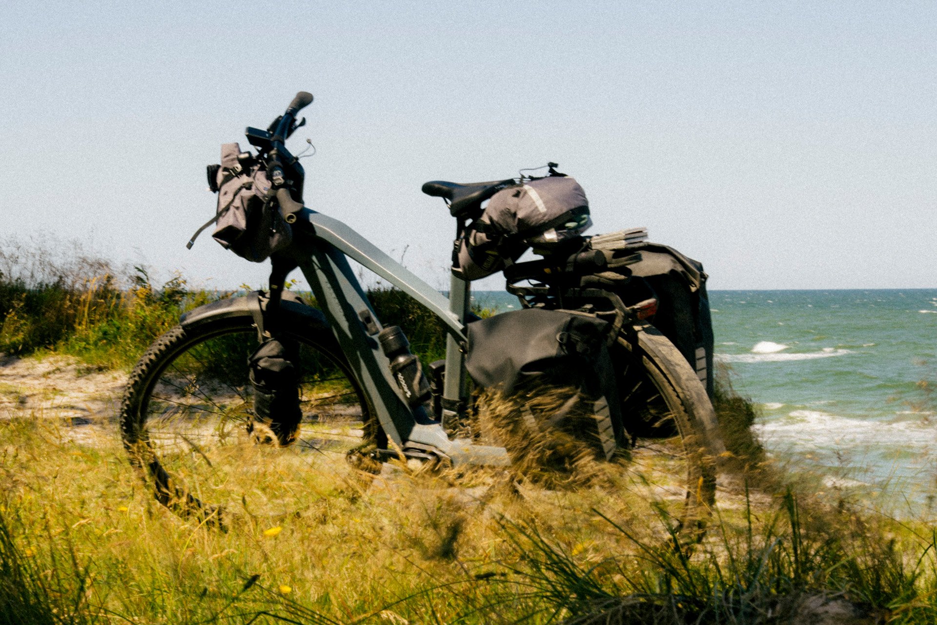 A loaded touring bicycle with panniers and bags is parked on grassy ground beside a serene, expansive ocean under a clear blue sky.