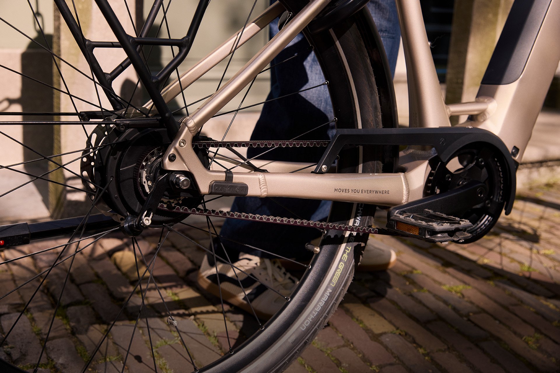 Nabenschaltungen haben in vielen Fällen Vorteile gegenüber Kettenschaltungen. Close-up of a bicycle's rear wheel and drivetrain, showing the belt drive system and pedal on a sunlit brick path.