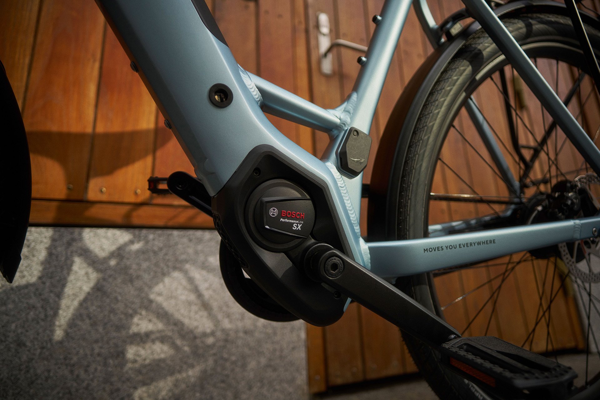 Close-up of a blue electric bicycle, showcasing the Bosch motor near the pedal, with a wooden door in the background.