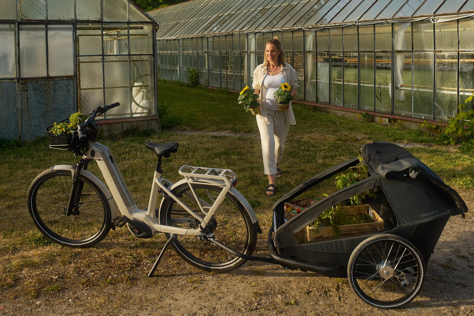 A woman walks near greenhouses, holding potted plants. A bicycle with a plant-filled trailer is parked on the grass.