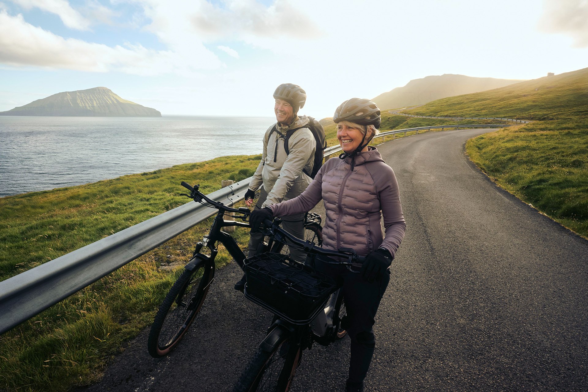 Two cyclists pause on a scenic coastal road, smiling and enjoying the view of the ocean and distant mountains under a clear sky.