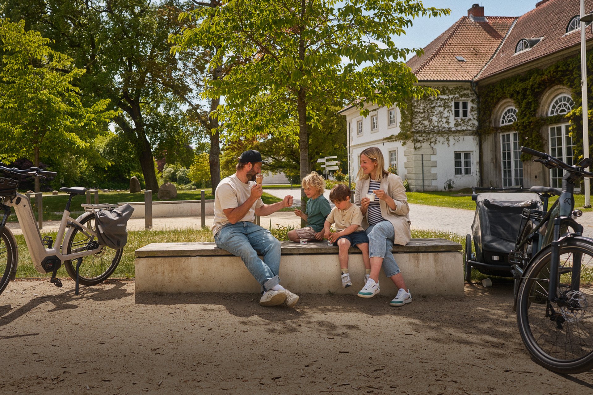 Mit der richtigen Vorbereitung bringt eine Radtour mit Kindern viel Spaß. A family of four enjoys snacks on a bench, surrounded by parked bicycles, with a house and lush trees in the background.