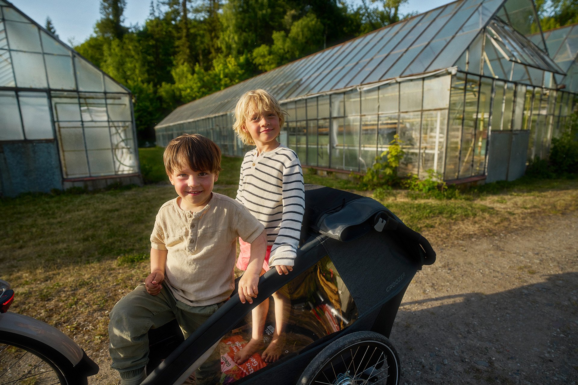 Two children smiling in a bike trailer, parked on a dirt path near greenhouses, with trees and sunlight in the background.