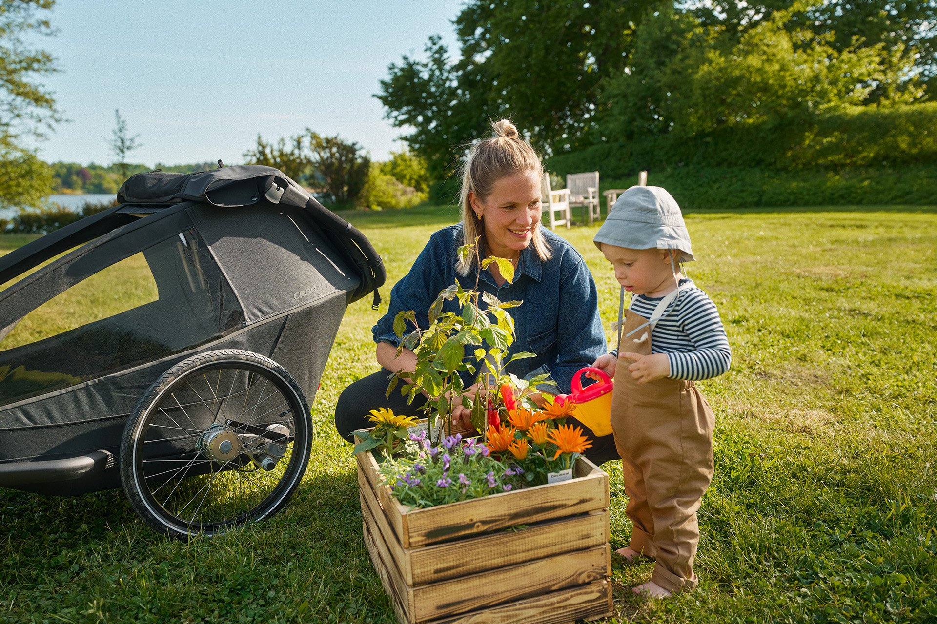A woman and a child gardening together, with a stroller nearby, on a sunny day in a grassy park.
