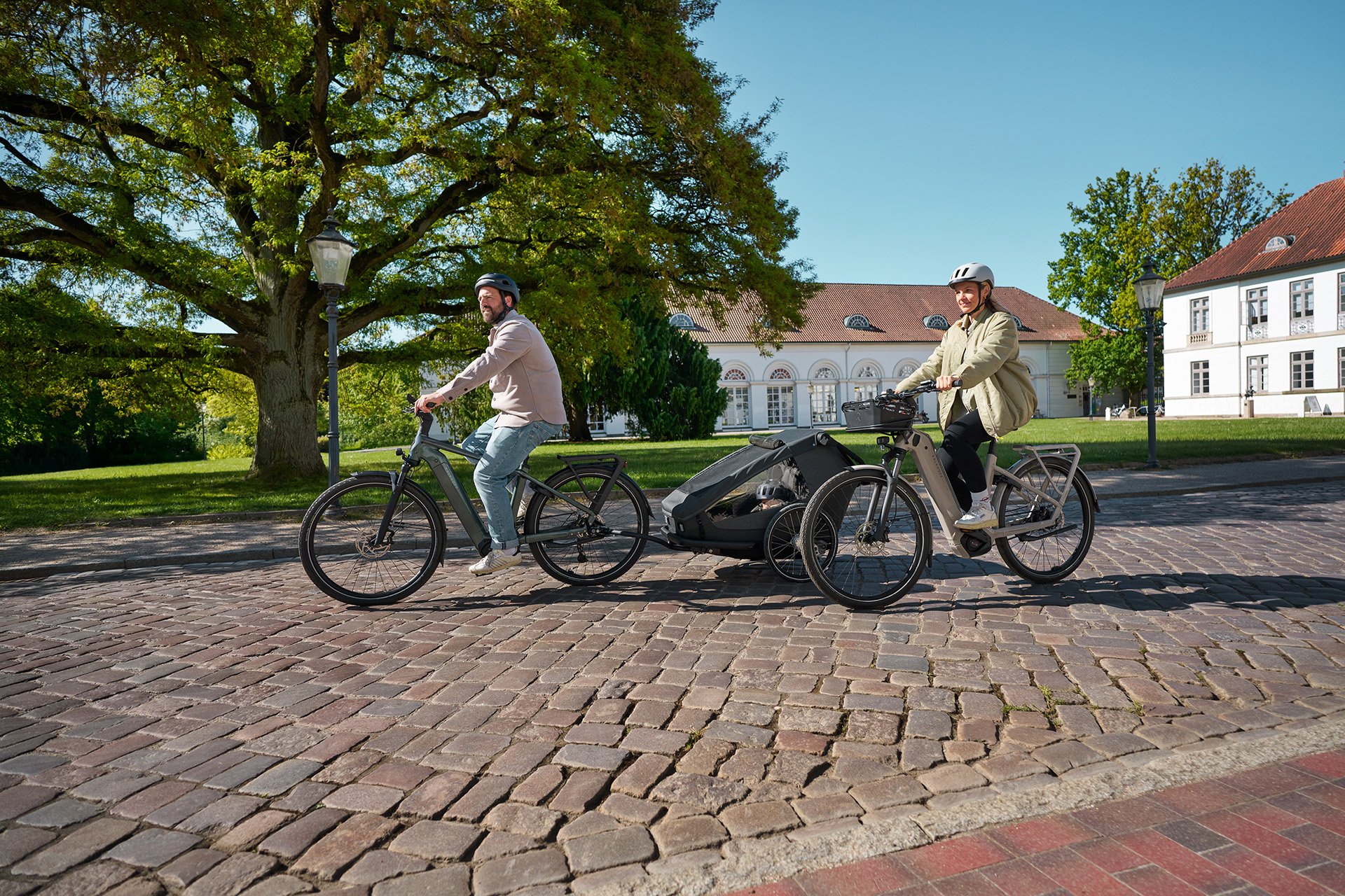 Two people riding e-bikes with a child trailer on a sunny cobblestone path, surrounded by trees and historic buildings.