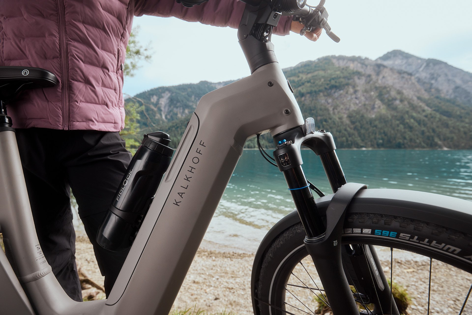 Person in a pink jacket standing with a Kalkhoff e-bike by a scenic lake, with mountains in the background.
