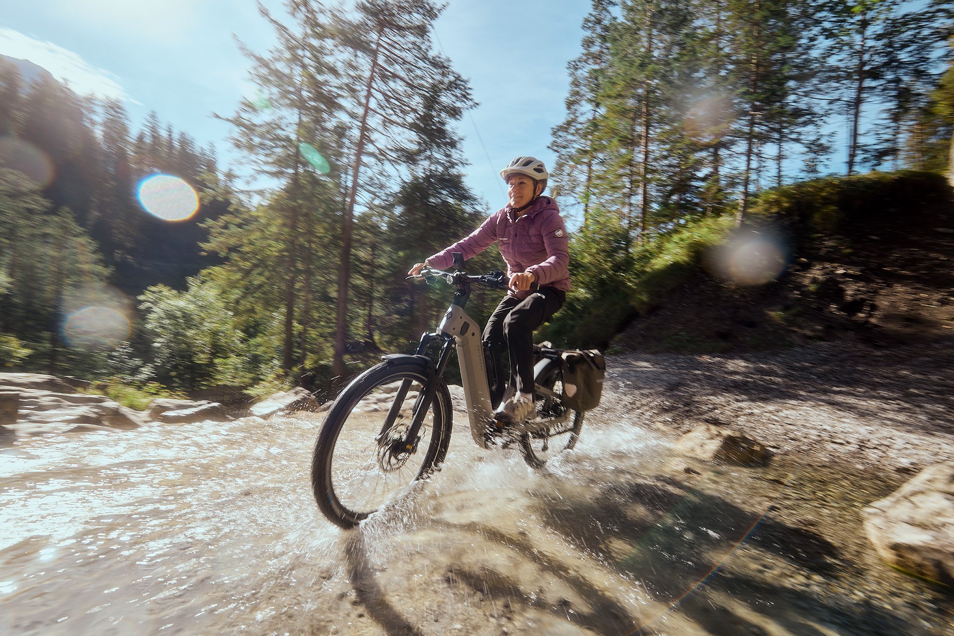 Person riding an electric bike through a shallow stream in a forested area on a sunny day, wearing a helmet and jacket.
