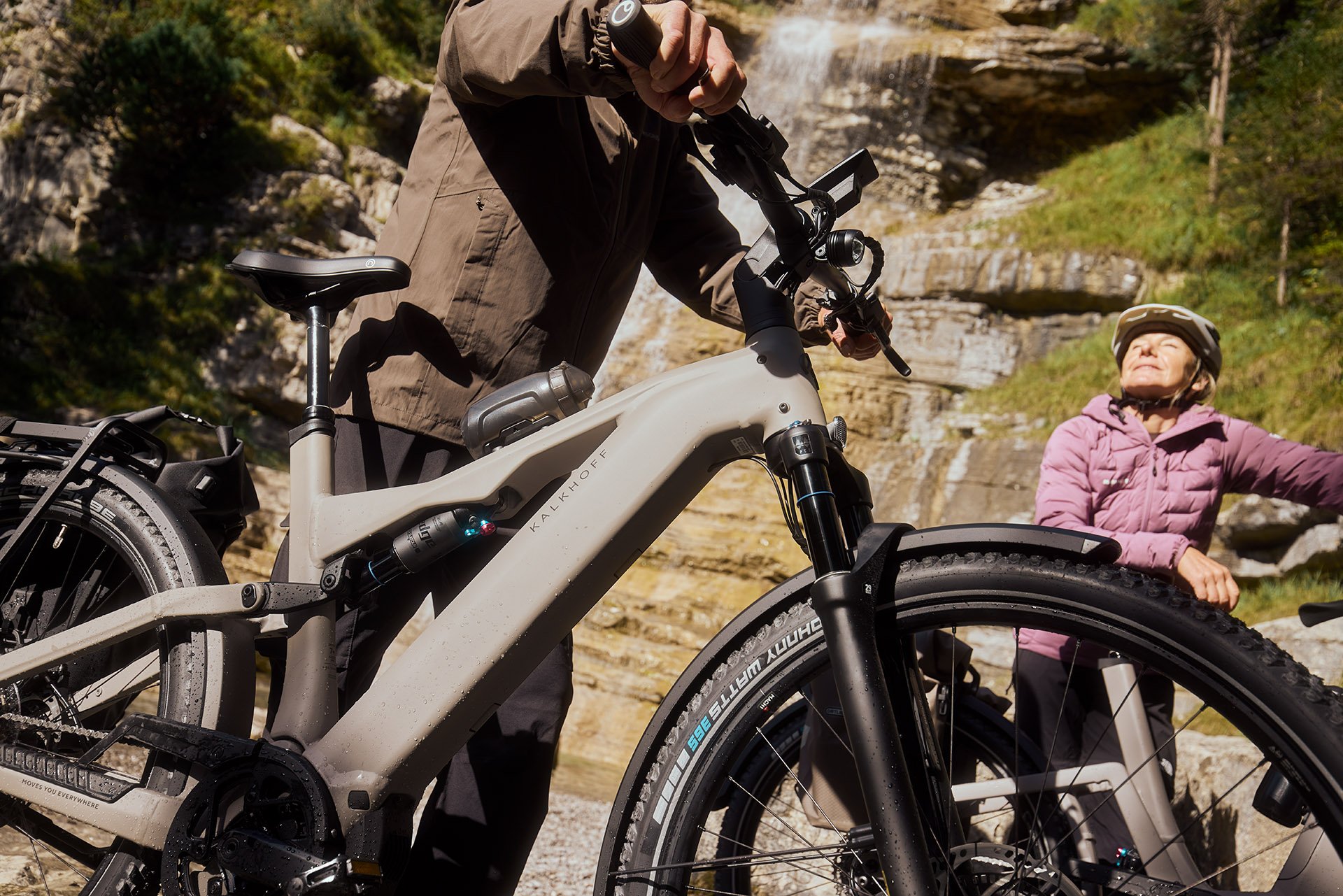 Two people with bicycles near a waterfall. One holds a bike, the other wears a helmet and looks up. Rocky and green landscape in the background.