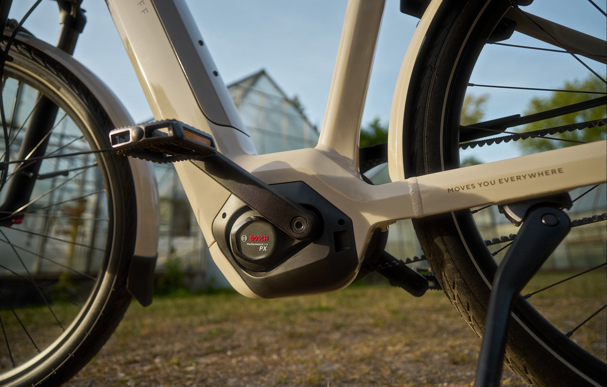 Close-up of an e-bike's pedal and motor system, showing the Bosch logo, with greenhouses blurred in the background.