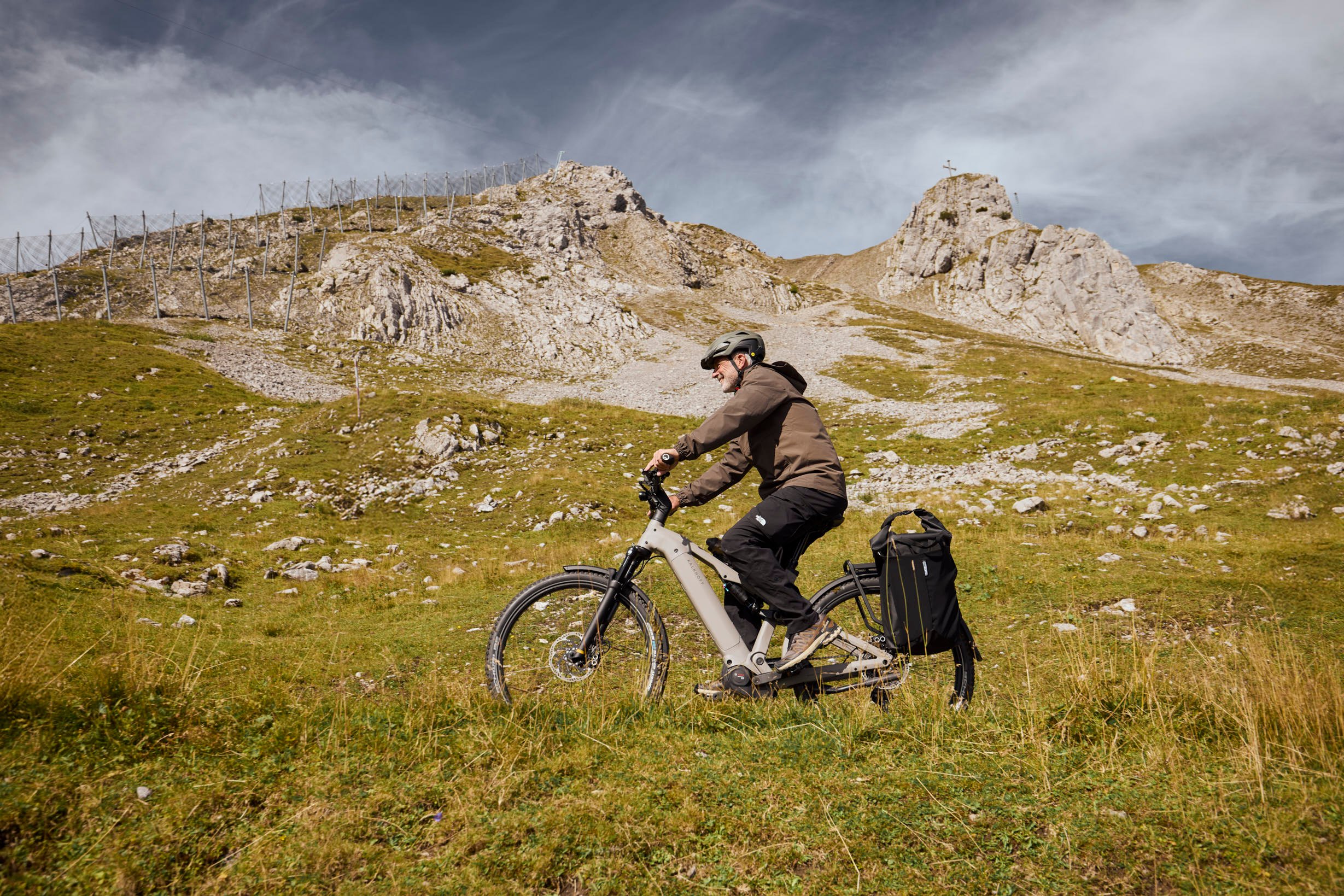 Sattel und Sattelstütze müssen für eine reibungslose Fahrt zusammenpassen. A cyclist rides an electric bike up a grassy mountain path, wearing a helmet and jacket, with rugged rocky hills in the background under a cloudy sky.