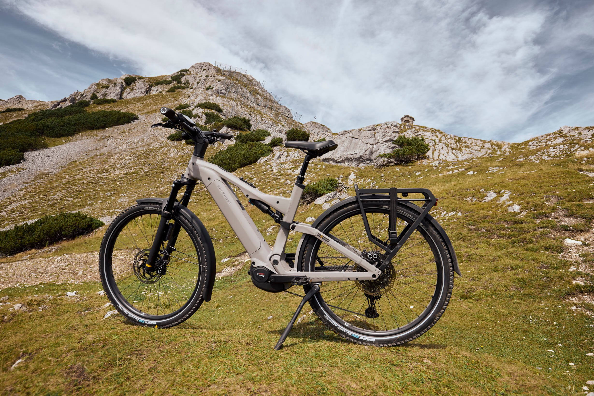 Der folgende Leitfaden hilft dir, den perfekten E-Bike Sattel zu finden. A gray e-bike with black tires and rear rack stands on a grassy hillside with rocky terrain under a partly cloudy sky.