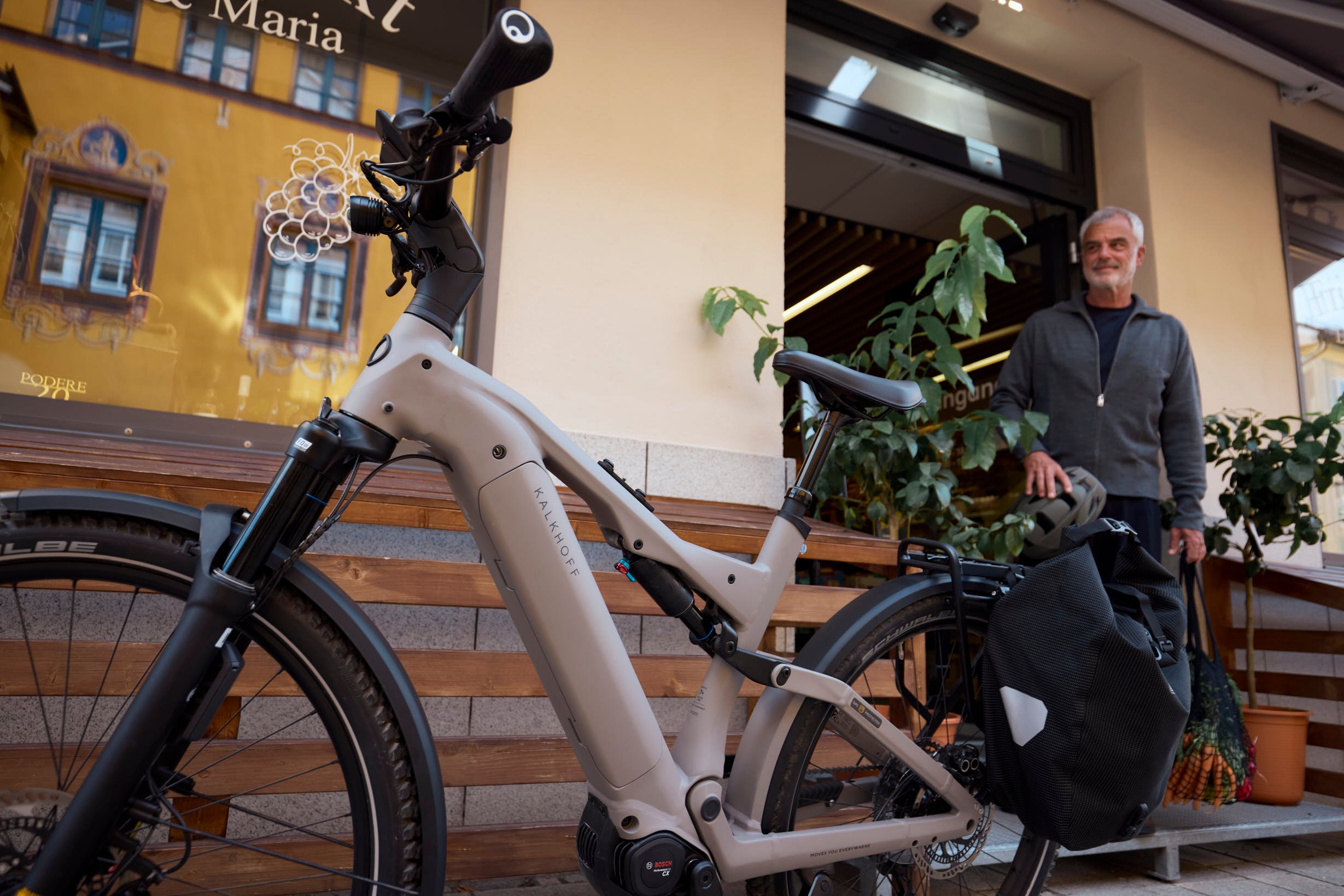 Der Einsatzzweck hat einen großen Einfluss auf die Sattelwahl. Electric bike with a rear bag parked near a shop entrance; a man with a helmet and groceries stands nearby.