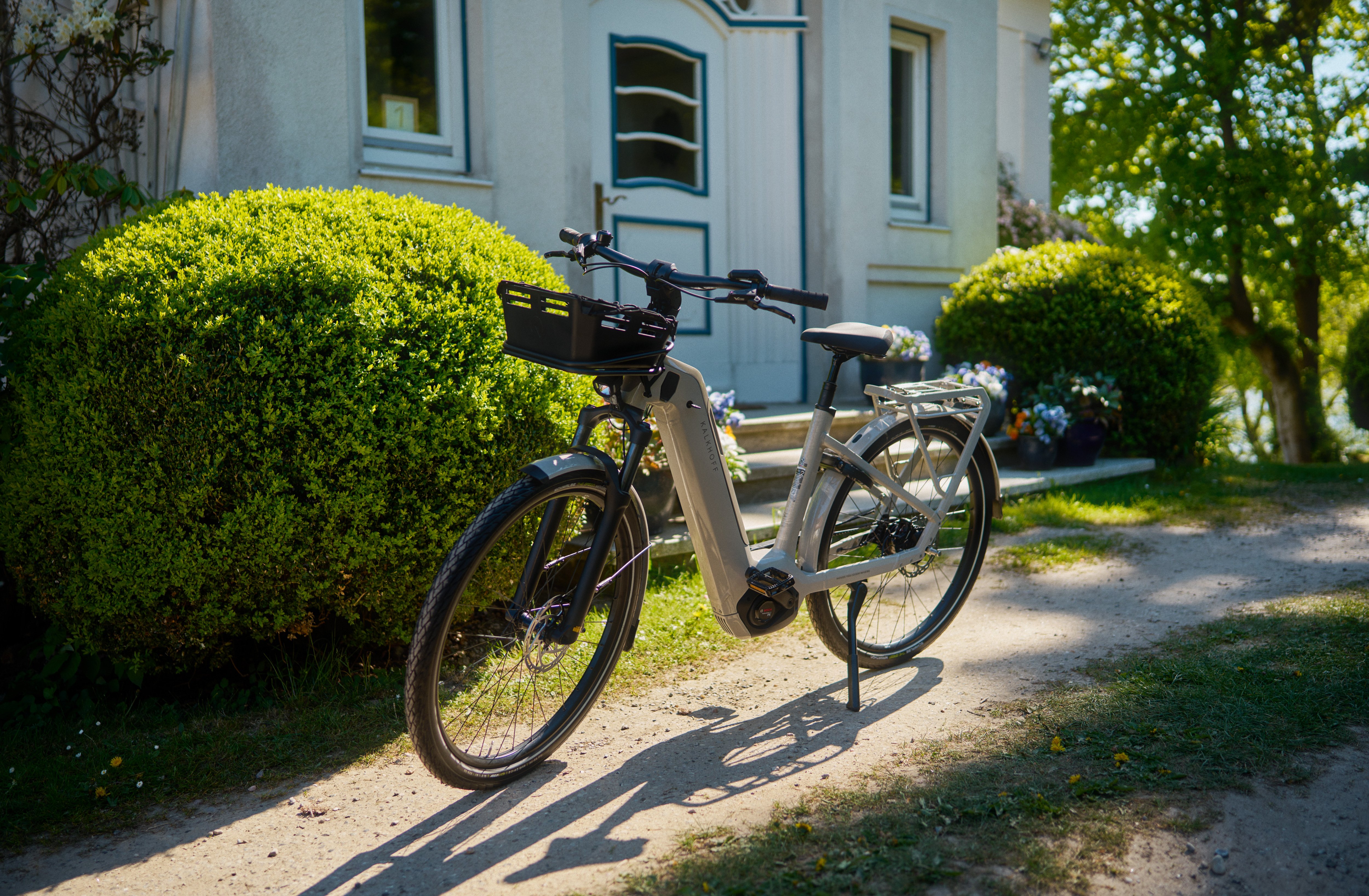 Ein modernes Kalkhoff Image 5, das auf einem sonnendurchfluteten Weg vor einem Haus mit Hecken und viel Grün geparkt ist.